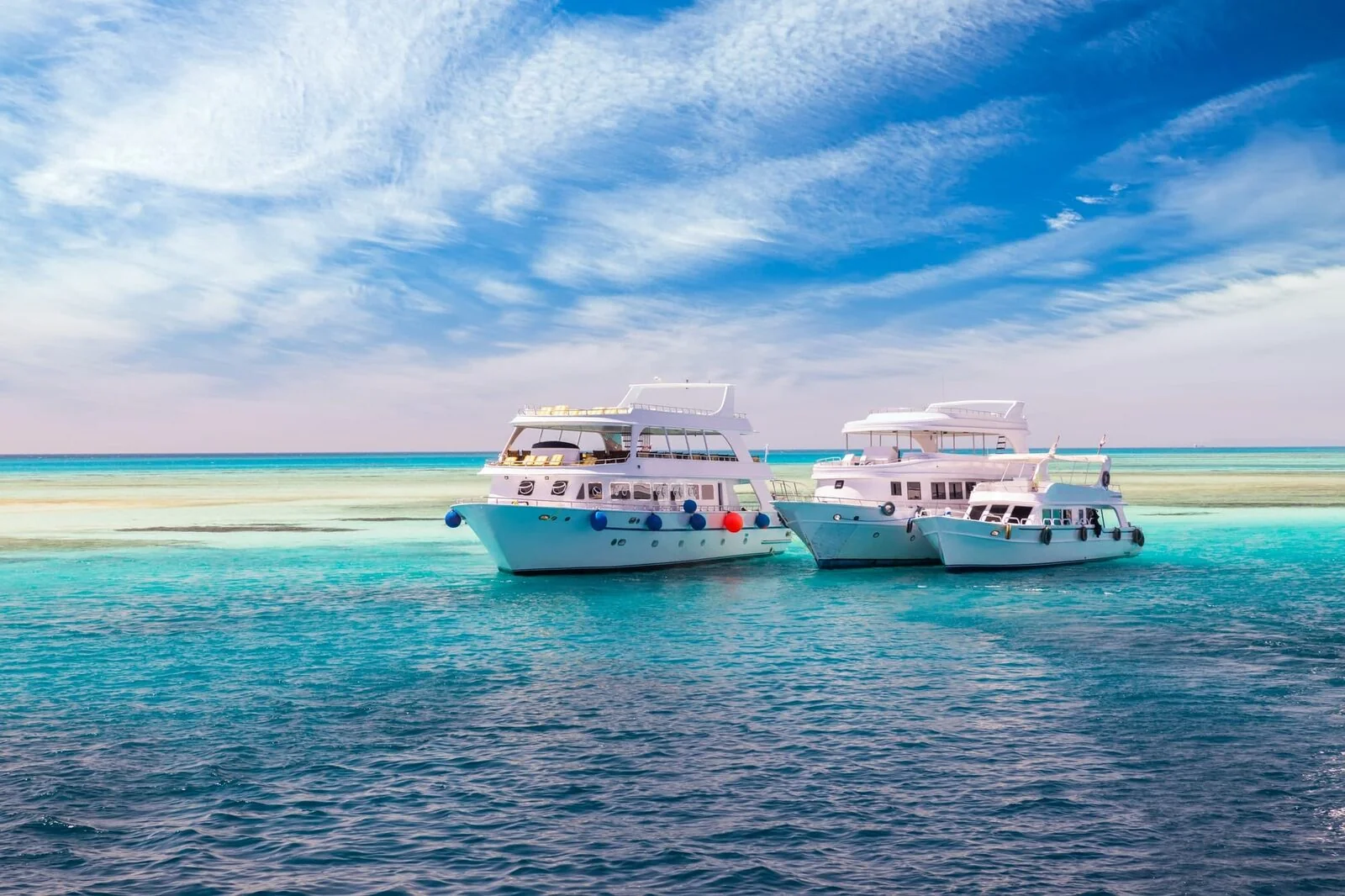 Three white tour boats anchored side by side in turquoise water near a sandbar under a partly cloudy sky.