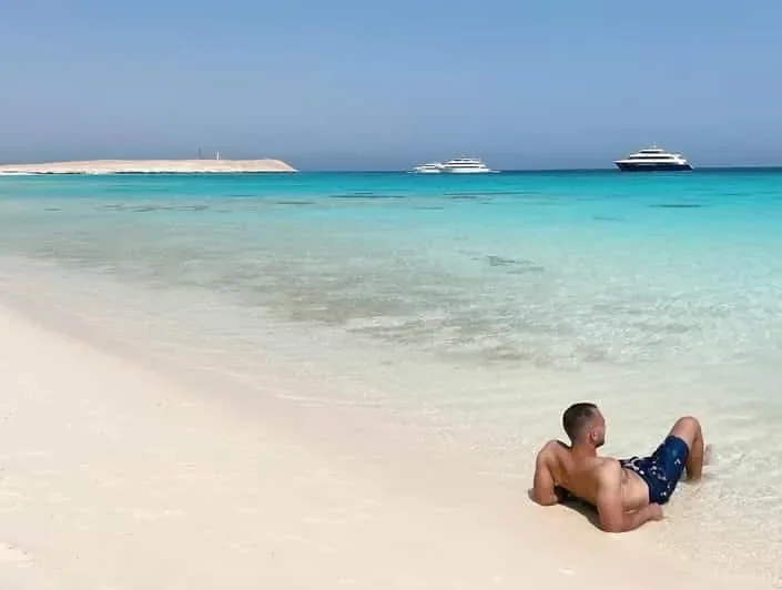 A man lying in shallow turquoise water on a white sand beach with tour boats moored offshore.