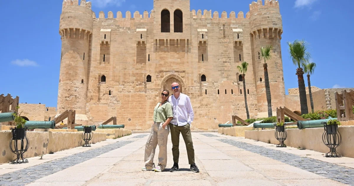 A couple posing together on the stone walkway leading to the entrance of the Citadel of Qaitbay, flanked by old cannons and palm trees.