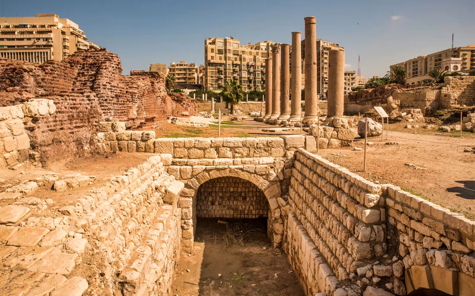 Ancient stone ruins featuring a vaulted brick archway and a row of tall standing columns at an archaeological site in Alexandria.