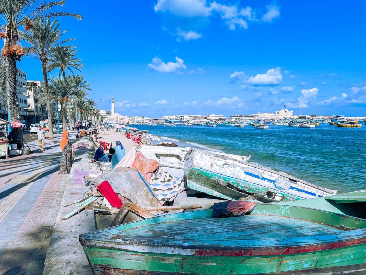 Weathered wooden fishing boats resting on the Corniche promenade beside the harbor, with palm trees and the Citadel of Qaitbay in the background.