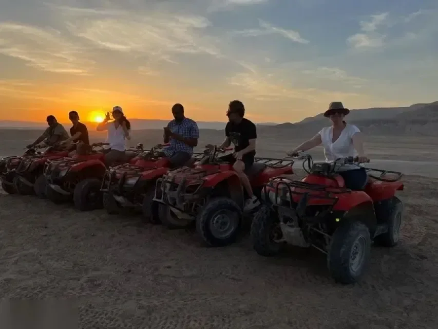 Six riders on red ATV quad bikes lined up in the desert as the sun sets behind distant hills.