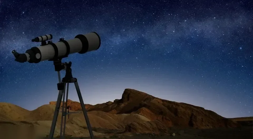 A telescope on a tripod is silhouetted against a starry night sky above rocky desert hills.