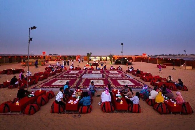 Rows of red cushioned low tables are set up for groups at an outdoor desert dinner venue at dusk.