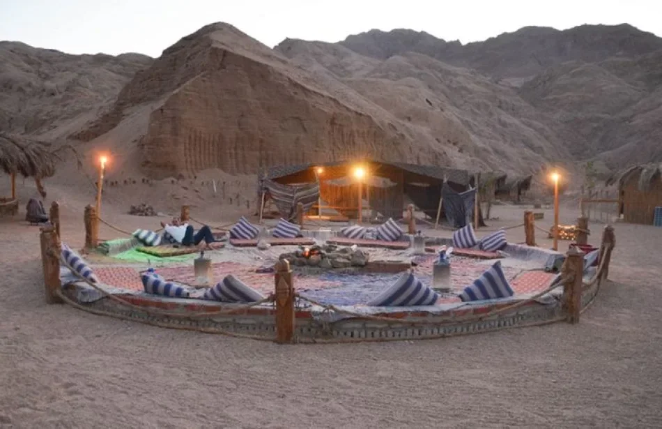 Two women on camels hold hands and smile at each other with mountains behind them at sunset.