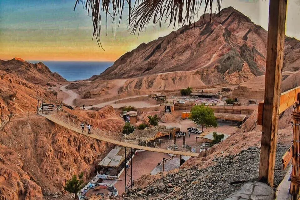 A rope suspension bridge crossing a rugged desert canyon near Dahab, with a small Bedouin camp and the Red Sea visible beyond.
