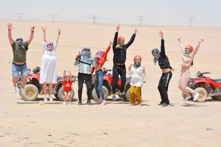 A group of tourists in ATV gear jump and cheer in the desert next to a row of red quad bikes.
