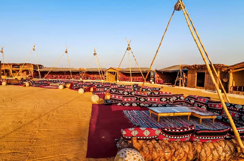 An empty Bedouin-style seating area with red patterned cushions under pyramid-frame canopies at a desert camp.