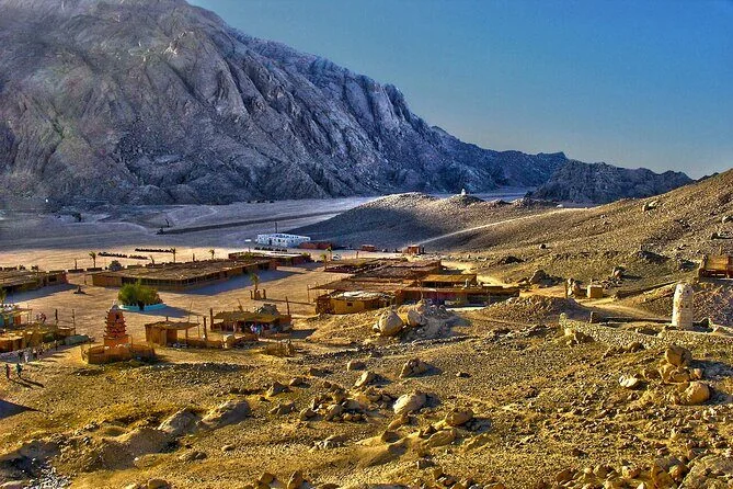 A man in a headscarf looks out over a Bedouin village of straw-roofed huts and resting camels below rocky mountains.