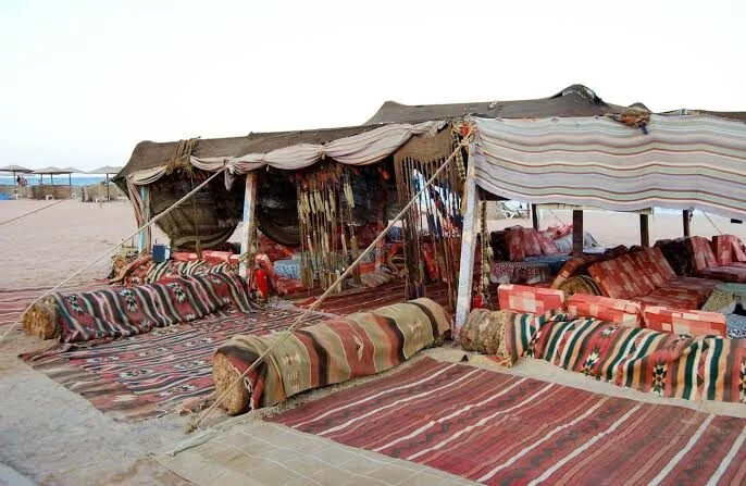 Elevated view of a desert Bedouin village compound with huts, courtyards, and parked vehicles beneath rocky mountains.