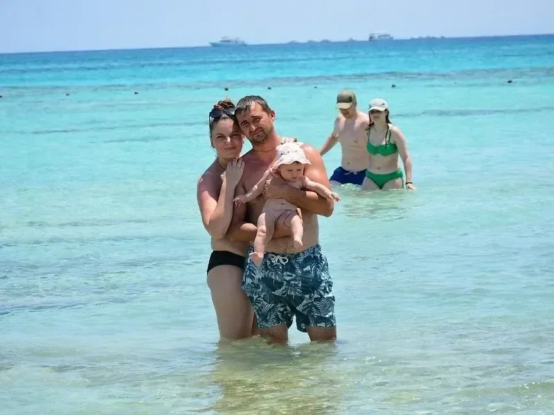 A family wades in shallow turquoise water, with the father holding a baby, while another couple stands in the background.