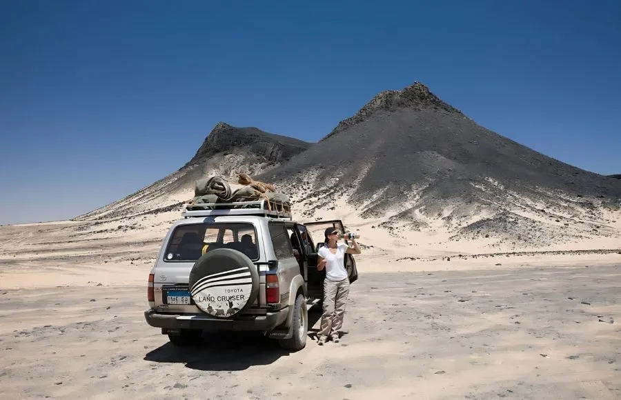 한 여성이 검은 사막 봉우리 앞에 주차된 은색 4x4 랜드크루저 옆에 서서 카메라로 사진을 찍고 있다.