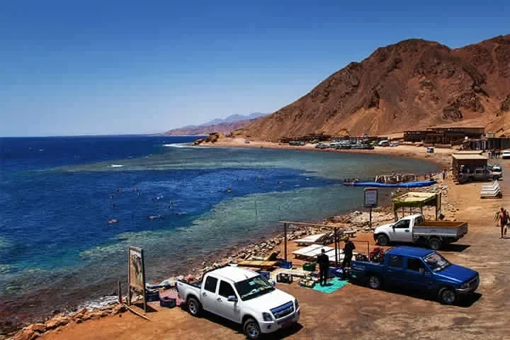 A coastal view of red desert mountains meeting a turquoise bay, with blue and white pickup trucks parked along the shoreline road.