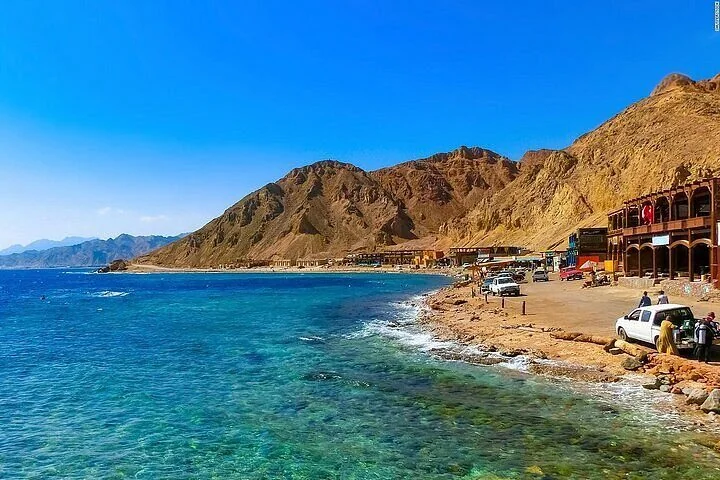 A wide coastal view of Dahab's shoreline with red mountains behind a row of buildings along the blue Red Sea water.