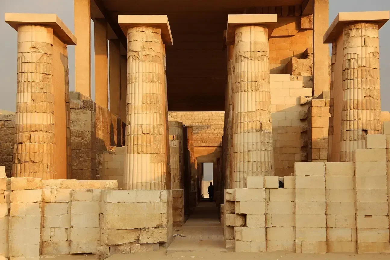 Rows of carved stone columns line an ancient temple corridor at Saqqara, with a person walking through it in the distance.