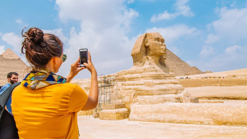 A woman photographs the Great Sphinx with her phone while a man stands beside her, pyramids visible in the background.