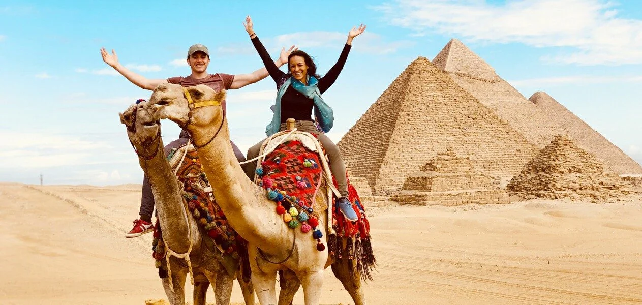 A couple rides camels with arms raised in celebration, with the Giza pyramids filling the background.
