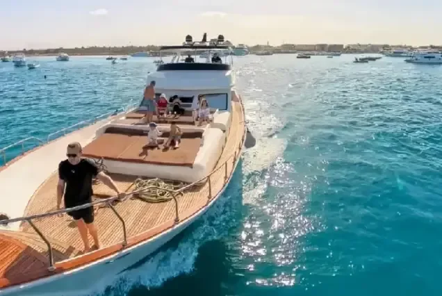 An aerial view of a yacht's bow deck where a small group relaxes on cushioned seating, with a harbor visible in the background.