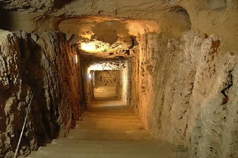 A torch-lit stone staircase descends through a narrow rock-cut corridor inside the Catacombs of Kom el Shoqafa.