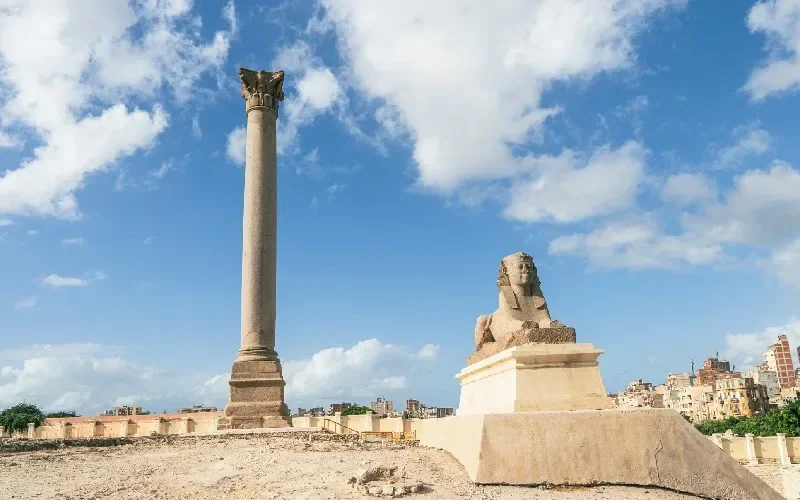 A tall Corinthian column known as Pompey's Pillar stands beside a seated sphinx statue with the city of Alexandria in the background.