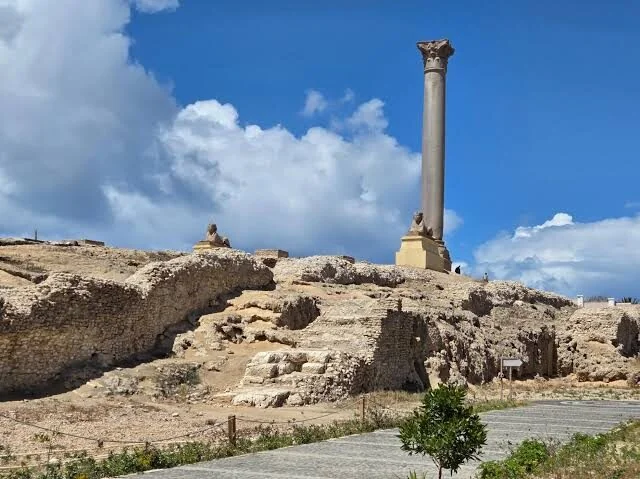 A wide view of the Pompey's Pillar archaeological site shows the column and a small sphinx statue atop excavated stone ruins, with a paved walkway in the foreground.