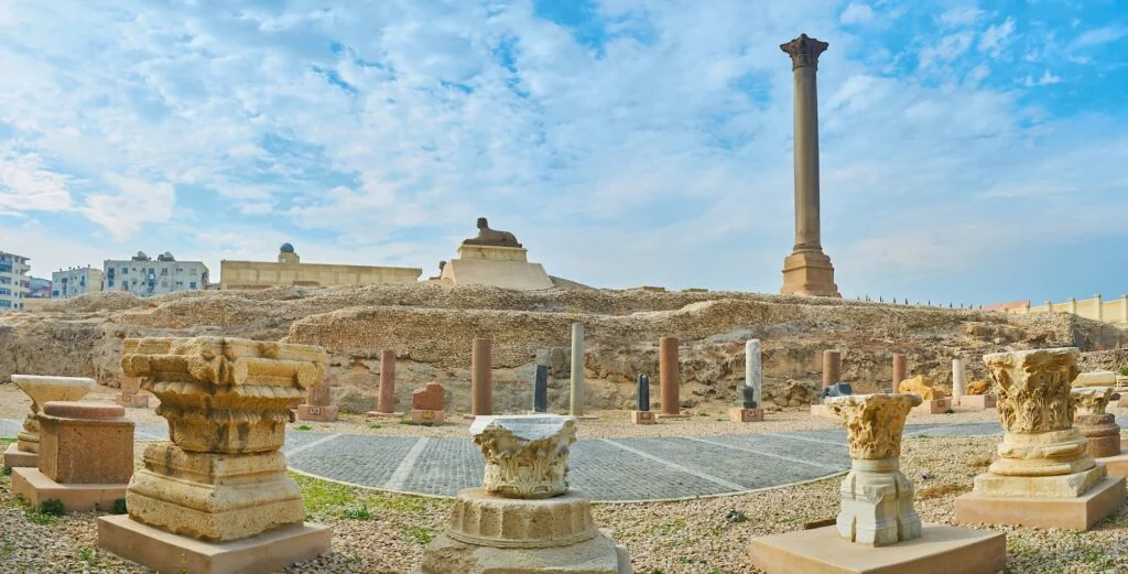 A panoramic view of the Pompey's Pillar grounds shows rows of ancient column capitals and bases arranged in the foreground, with the pillar and sphinx statue on a rise in the background and city buildings beyond.