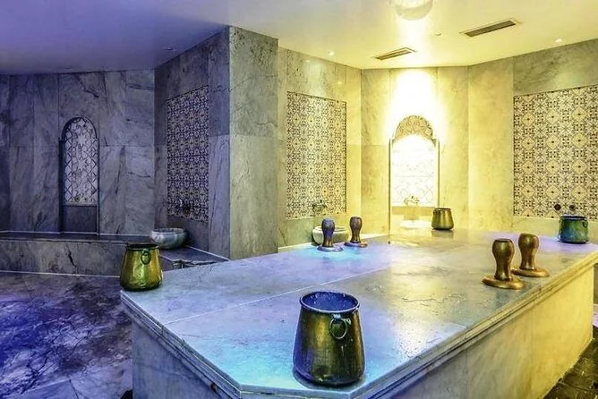 A marble hammam washing room with arched wall niches, patterned tile inlays, and several copper wash bowls (tas) sitting on a marble platform.