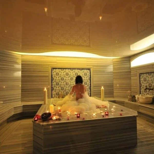 A woman sits on a marble platform covered in bath foam, seen from behind, surrounded by lit candles in a warmly lit hammam bath room.