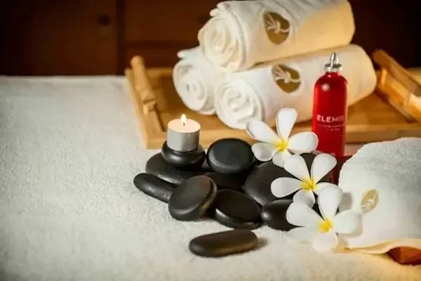 A spa still-life of rolled white towels, black hot stones, a lit candle, white frangipani flowers, and a red massage-oil bottle arranged on a light-colored towel.