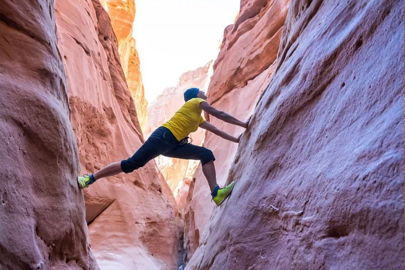 A person in a yellow shirt braces hands and feet against two narrow canyon walls, climbing between them above the ground.