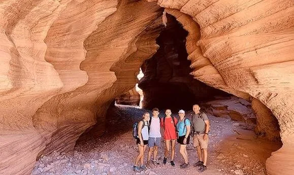 A group of five hikers with backpacks poses smiling inside a wide, cave-like opening in a reddish sandstone canyon.