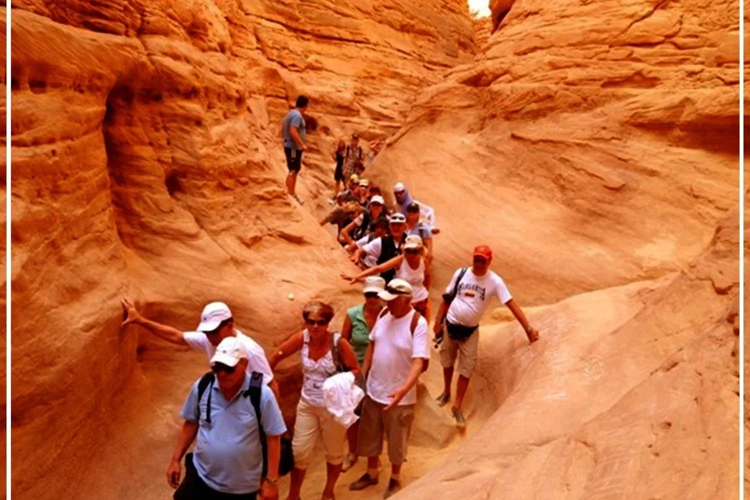 A long line of tourists in sun hats walks through a wide, sandy corridor between tall orange sandstone canyon walls.