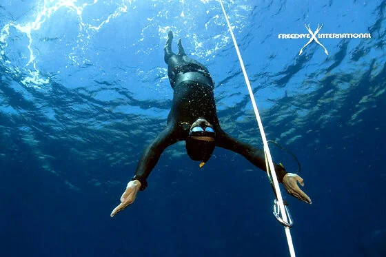 Freediver descends headfirst along a rope in deep blue water, trailing bubbles behind.