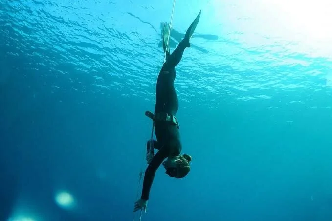 Freediver hangs upside down just below the surface, fins pointed upward toward the light.