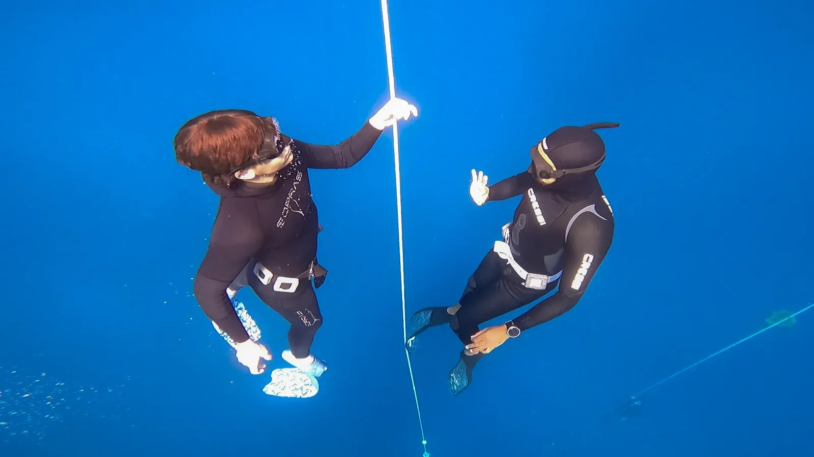 Two freedivers in black wetsuits face each other at depth, one holding onto the guide line.