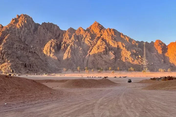 Wide desert valley with jagged orange-lit mountains, palm trees, and a distant quad bike crossing the sand.