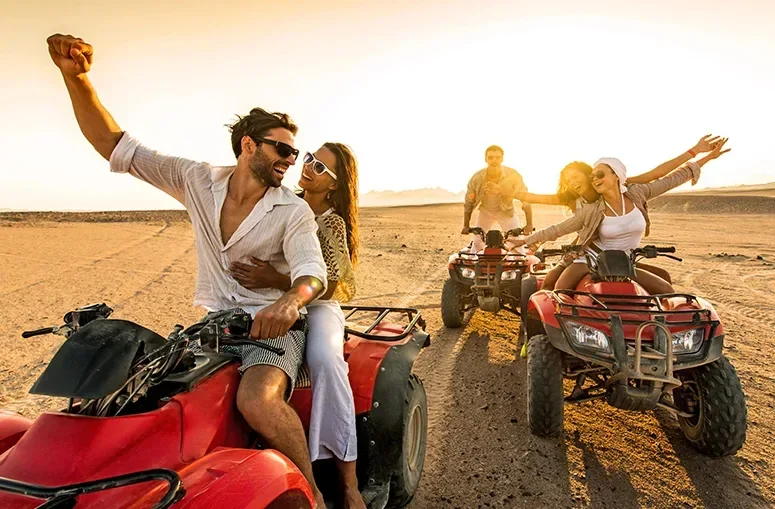 A couple poses cheerfully on a red quad bike with friends nearby during golden hour in the desert.