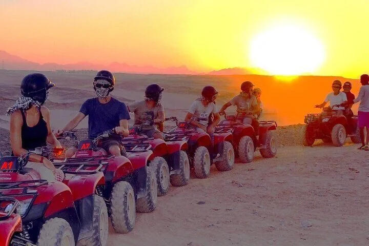 A line of quad bike riders in helmets and scarves pauses against a huge glowing orange sunset.