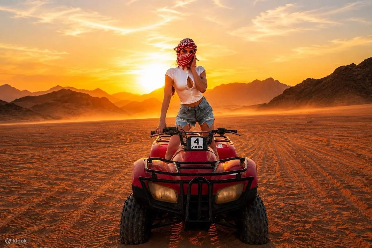 A woman in a red-and-white headscarf stands on a quad bike against a fiery desert sunset.