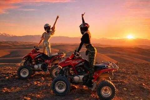 Two women in helmets pose with arms raised on their quad bikes against a colorful sunset sky.