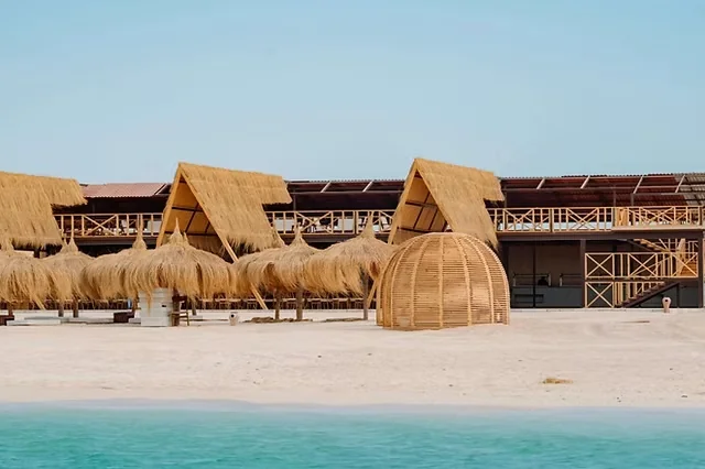 Row of thatched-roof beach cabanas and a domed straw hut on white sand beside turquoise water.