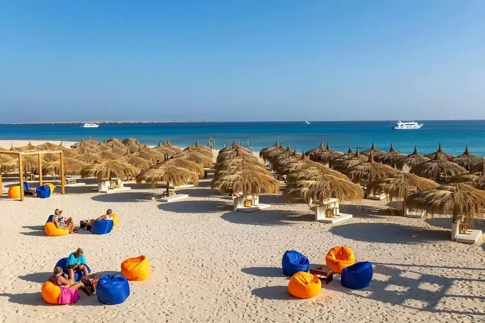 Beach club with rows of straw umbrellas and colorful bean bag loungers on the sand, boats visible offshore.