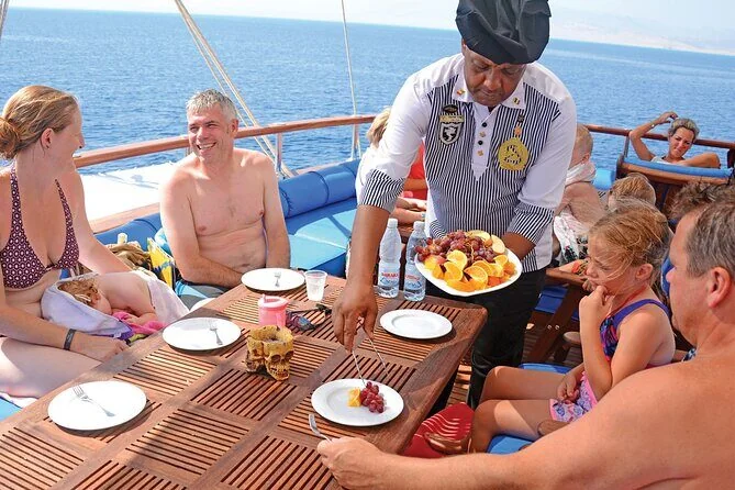 Uniformed steward serving a platter of fresh fruit to a family seated at an outdoor dining table on the yacht.