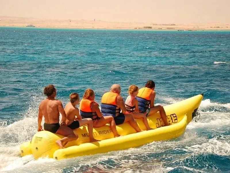 Family of six in orange and blue life vests riding a yellow banana boat towed across the sea, desert coastline behind them.
