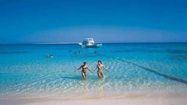 Two women in bikinis holding hands and walking through shallow turquoise water toward the beach, with a boat and swimmers in the background.