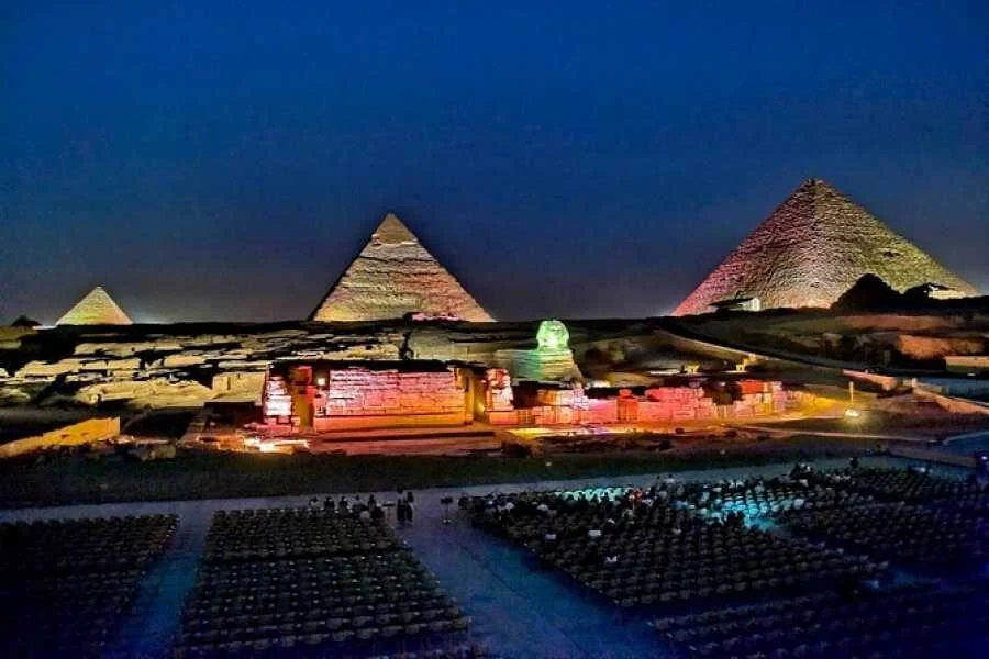 Night view of the illuminated Giza pyramids and Sphinx with rows of empty audience seating in front.