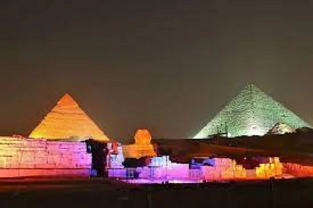 Night view of the illuminated Giza pyramids and Sphinx with rows of empty audience seating in front.