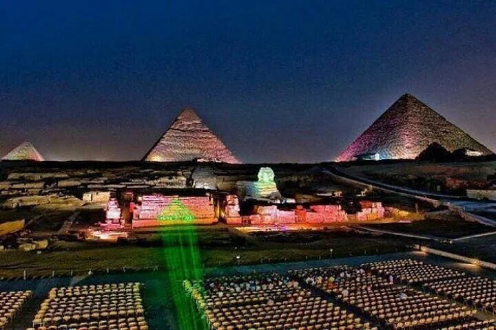 Close-up night view of two pyramids lit in orange and green with the Sphinx illuminated between them.