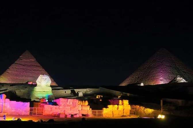 Aerial night view of the pyramids, Sphinx, and rows of audience seating with a green laser beam crossing the scene.
