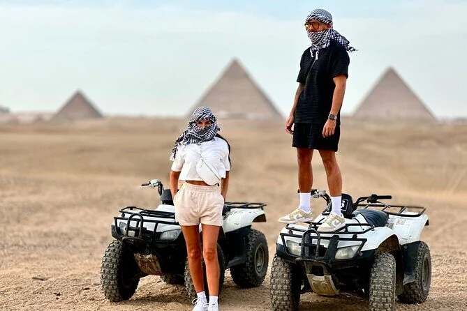 Two riders in checkered headscarves stand on parked quad bikes in the desert with pyramids visible in the distant background.